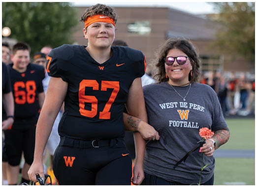 An adult wearing a football uniform with the number 67 stands arm-in-arm with another adult wearing sunglasses and a West De Pere Football t-shirt while holding a flower on a sports field. Description generated by AI