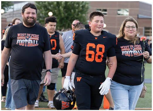 A person in a football uniform with the number 68 walks on a field holding a helmet and is accompanied by two adults wearing matching West De Pere Football shirts. Description generated by AI