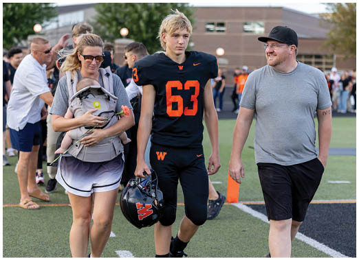 An adult carrying a newborn in a baby carrier walks alongside a person in a black football uniform holding a helmet and another adult in casual clothing on a sports field. Description generated by AI