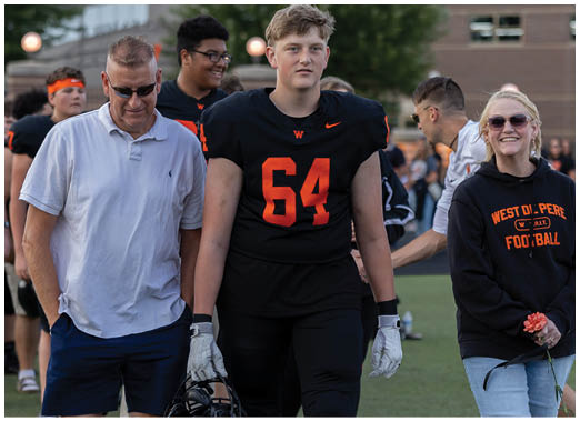 An adult in a white polo shirt and shorts walks beside a person in a black football uniform holding a helmet, while another person wearing sunglasses and a black sweatshirt with football text holds a flower on a sports field. Description generated by AI