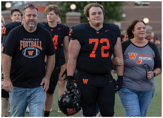 A group of people on a football field includes a person in a black football uniform holding a helmet and two others wearing casual clothing with football-themed shirts. Description generated by AI