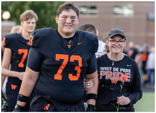 A person wearing a black football uniform with the number 73 is smiling and walking arm-in-arm with another person wearing glasses and a black shirt that says “West De Pere Pride" while another person in a similar football uniform stands in the background. Description generated by AI