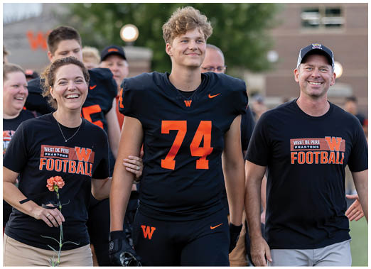 A person wearing a football uniform with the number 74 is walking arm-in-arm with two people wearing matching football-themed t-shirts, all smiling outdoors. Description generated by AI
