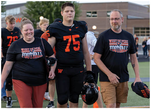 A person in a football uniform with the number 75 walks on a field holding hands with two adults wearing matching West De Pere Football t-shirts, with other people and a building in the background. Description generated by AI
