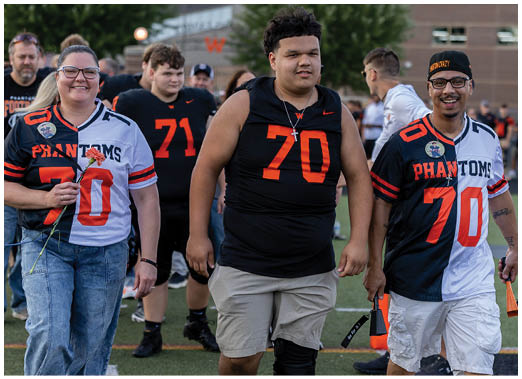 Three adults wearing sports jerseys with the number 70 walk together on a field, smiling and holding flowers and flags. Description generated by AI