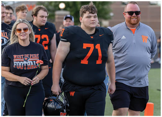 A group of people walk together on a sports field with one person in a black football uniform holding a helmet and others wearing casual sportswear, including a shirt that says “West De Pere Football." Description generated by AI