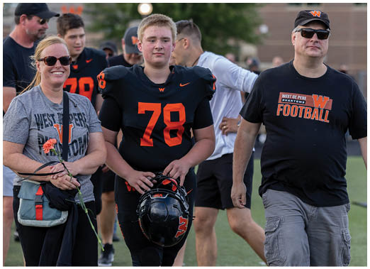 A person in a football uniform holding a helmet stands between two people wearing casual clothing and smiling on a sports field. Description generated by AI