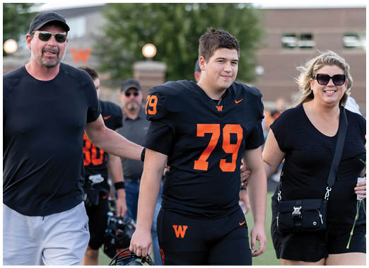 A person wearing a black football uniform with the number 79 is walking on a field while holding hands with two adults dressed in casual clothing. Description generated by AI