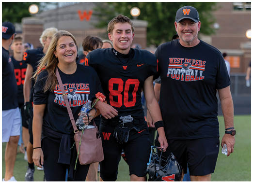 Three adults wearing matching West De Pere Football shirts walk together on a sports field with one adult in a football uniform holding a helmet and smiling. Description generated by AI