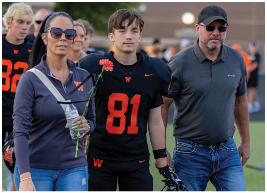 An adult wearing sunglasses and casual clothing stands next to a person in a football uniform holding a flower, accompanied by another adult in casual clothing and a cap, with a sports field and other people in the background. Description generated by AI