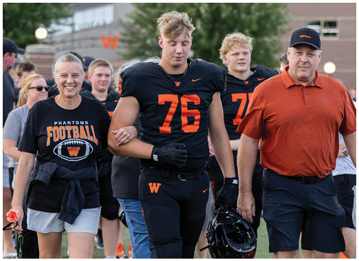 A person wearing a black football uniform with the number 76 walks arm-in-arm with two adults, one in a black football t-shirt and shorts holding a flower, and the other in an orange polo and dark shorts, with other people and a building in the background. Description generated by AI