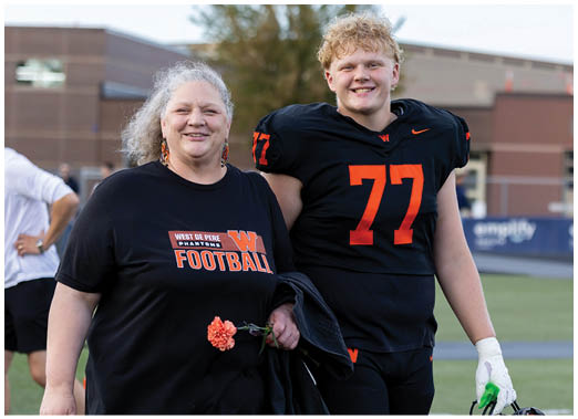 A person wearing a black football jersey with the number 77 stands next to another person in a black West De Pere Panthers football t-shirt holding a flower on a sports field. Description generated by AI