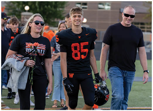 A person in a black football uniform with the number 85 walks on a field holding a helmet, accompanied by two adults wearing casual clothing and sunglasses, with other people and a building in the background. Description generated by AI