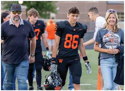 A person in a black and orange football uniform holding a helmet walks on a field accompanied by three people, one wearing a dark polo and cap, another in a gray sports shirt, and a third in a black and orange jersey. Description generated by AI