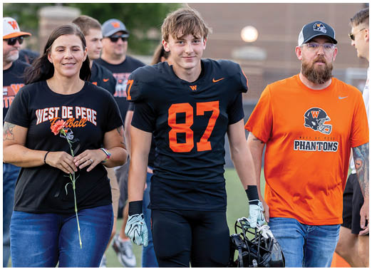 An adult in a football uniform walks on a field holding a helmet, accompanied by two adults wearing casual clothing with team logos, one holding a flower. Description generated by AI