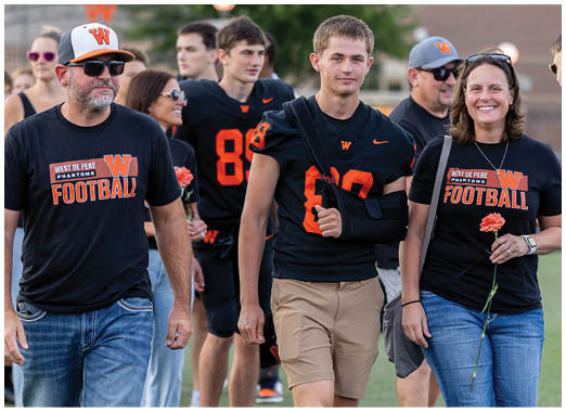 A group of people wearing West De Pere Phantoms football shirts and jerseys walk together outdoors, with one person holding a flower and another wearing an arm sling. Description generated by AI
