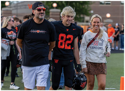 Three people stand together on a sports field, with one wearing a black football uniform holding a helmet, and the others dressed casually, all smiling. Description generated by AI