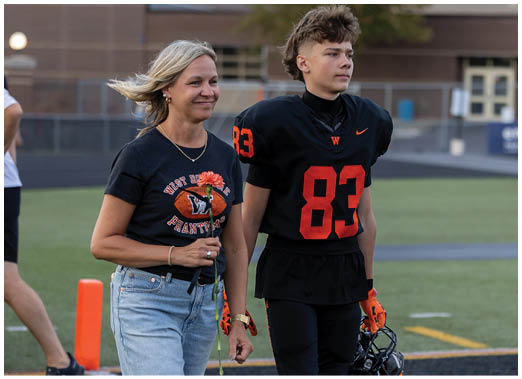An adult wearing a black t-shirt and jeans walks beside a person in a black football uniform holding a helmet on a sports field. Description generated by AI