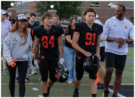 Two people in black football uniforms holding helmets walk alongside two people in casual sportswear on a football field with others in the background. Description generated by AI