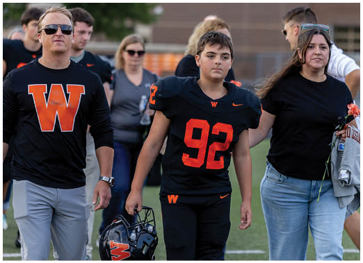 A person wearing a football uniform with the number 92 walks on a field holding a helmet, accompanied by two adults wearing casual clothing with a large orange W on their shirts. Description generated by AI