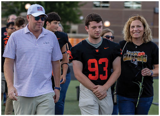 Three adults walk together on a sports field with one wearing a football jersey and another holding a flower, while other people are visible in the background. Description generated by AI