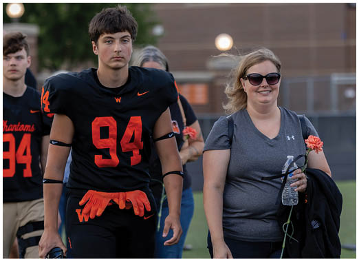 An adult wearing a black football uniform with the number 94 walks beside another adult in casual athletic clothing holding a water bottle and a jacket, both outdoors on a sports field. Description generated by AI
