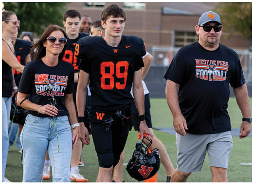 Three people stand together on a sports field, with one wearing a black football uniform holding a helmet, and the others dressed casually, all smiling. Description generated by AI
