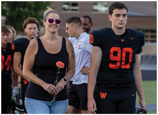 An adult wearing sunglasses and casual clothes holds a flower while standing among people in black sports uniforms with orange numbers on a field. Description generated by AI