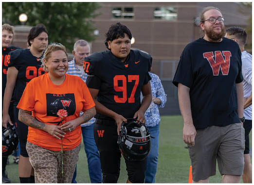 A group of people on a sports field includes one person in a football uniform holding a helmet, another person wearing an orange shirt holding a flower, and others walking alongside them. Description generated by AI