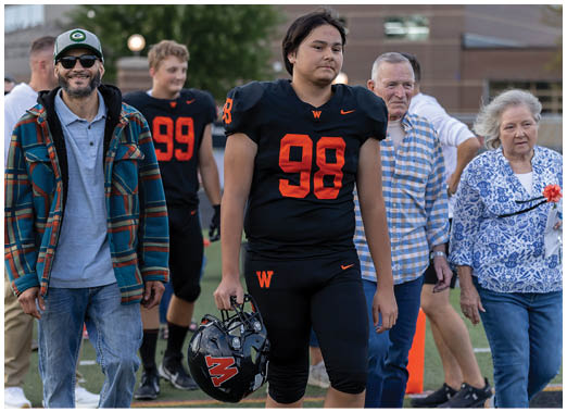 A person in a black football uniform holding a helmet walks on a field accompanied by four casually dressed people. Description generated by AI
