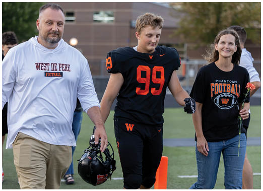 An adult in a white West De Pere Football shirt holds hands with a person in a black football uniform while walking alongside another adult wearing a black Phantoms Football shirt holding a flower on a sports field. Description generated by AI