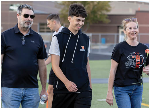 Three people walk together on a sports field with one holding a flower and another holding a water bottle, all appearing relaxed and smiling. Description generated by AI