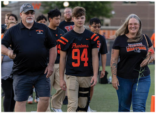 A group of people walk on a sports field with one person in a black and orange football jersey and others wearing casual clothing, including a person holding a flower. Description generated by AI