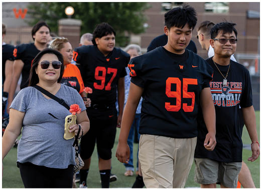 People wearing football uniforms and casual clothing are walking together on a sports field with trees and buildings in the background. Description generated by AI