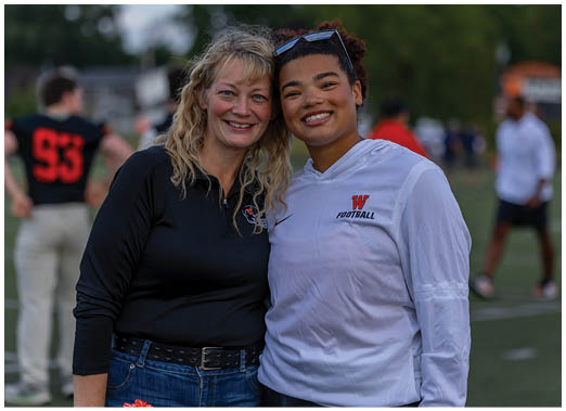 Two people smiling and standing close together on a sports field with others and trees in the background. Description generated by AI