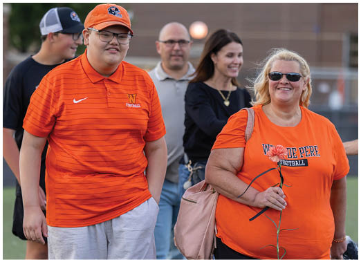 Two adults wearing orange sports shirts stand outdoors on a field, one holding a flower, with three other adults in the background smiling. Description generated by AI