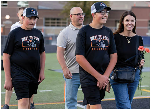 Two people wearing matching sports shirts and caps walk alongside two adults on a sports field, with one adult holding a flower and smiling. Description generated by AI