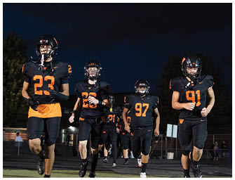 Five people wearing black and orange football uniforms and helmets are walking on a field at night. Description generated by AI