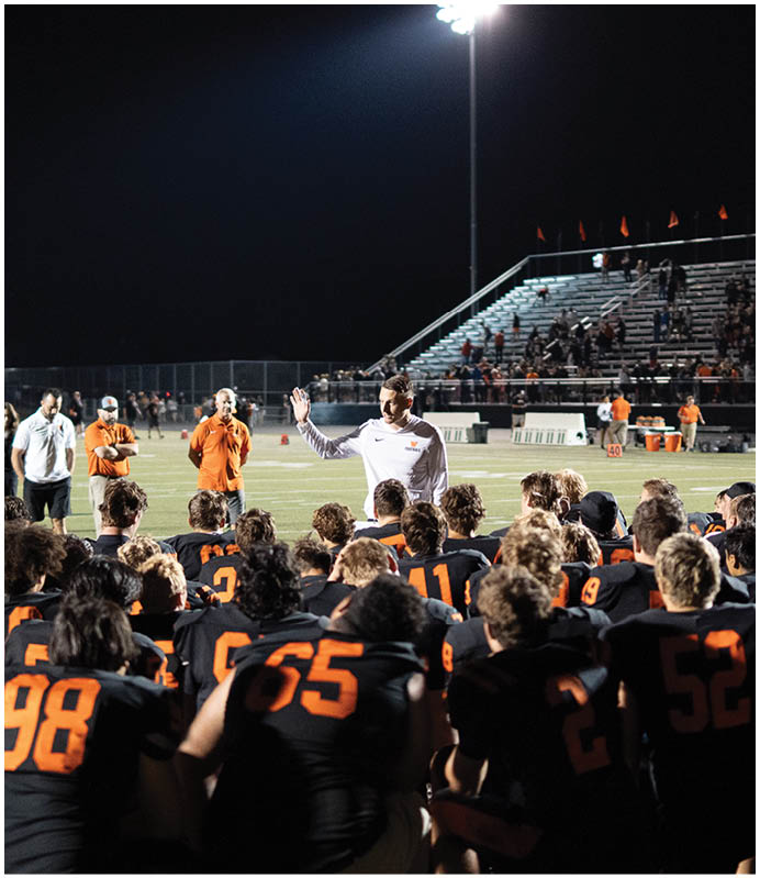 A group of people in black and orange sports uniforms are gathered on a football field at night while a person in white speaks to them with a raised hand. Description generated by AI