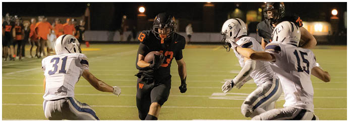 A group of people wearing football helmets and uniforms are actively playing on a lit field at night with others standing in the background. Description generated by AI