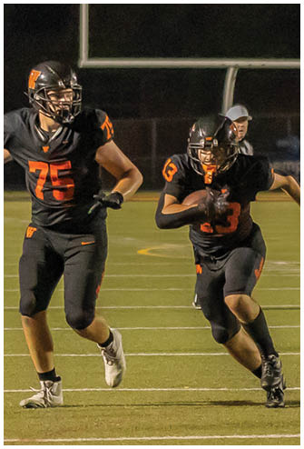 A person wearing a headset and holding a play sheet is coaching on a football field while a player in a black uniform with orange cleats walks nearby. Description generated by AI