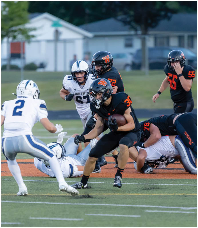 A group of people wearing black football uniforms with orange numbers and helmets are crouched in a formation on a grassy field. Description generated by AI