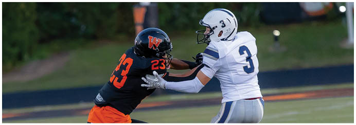 Two people in football uniforms are engaged in a play on a grassy field, with one wearing a black and orange uniform and the other in a white and gray uniform. Description generated by AI