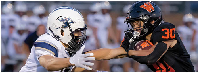 Two people in football uniforms are engaged in a play with one person holding the ball while the other reaches out to block or tackle. Description generated by AI