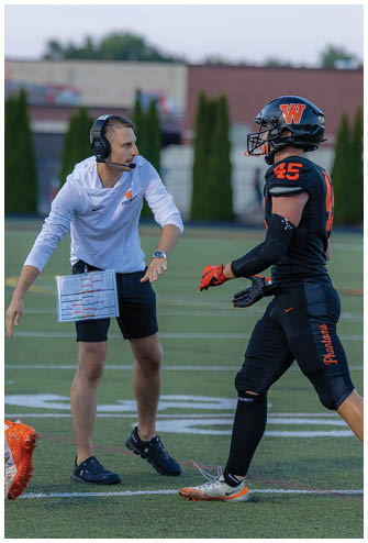 A person wearing a headset and holding a play sheet is coaching on a football field while a player in a black uniform with orange cleats walks nearby. Description generated by AI