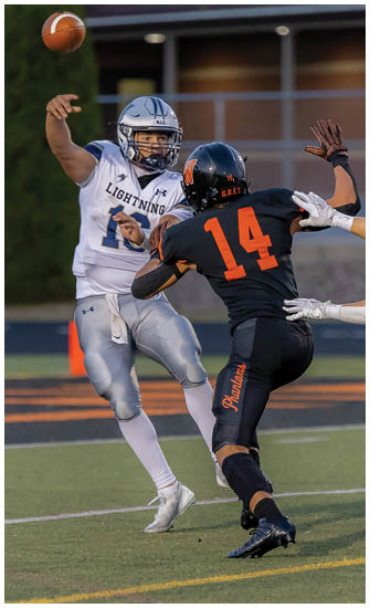 A person in a white and silver football uniform is throwing a football while another person in a black and orange uniform attempts to block the pass on a football field. Description generated by AI