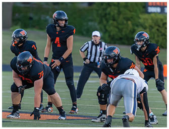 People in football uniforms are positioned on a field preparing for a play, with one person in a white uniform crouched opposite three people in black uniforms. Description generated by AI