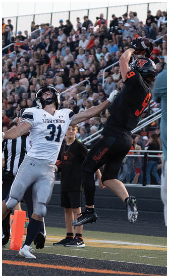 Two athletes in football uniforms compete to catch a ball near the end zone with a crowd and flags in the background. Description generated by AI