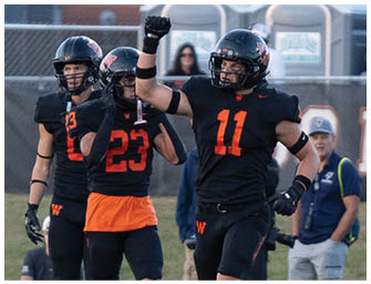 Three people in black and orange football uniforms celebrate on a field while a person in a white uniform walks away and several people stand near a fence in the background. Description generated by AI