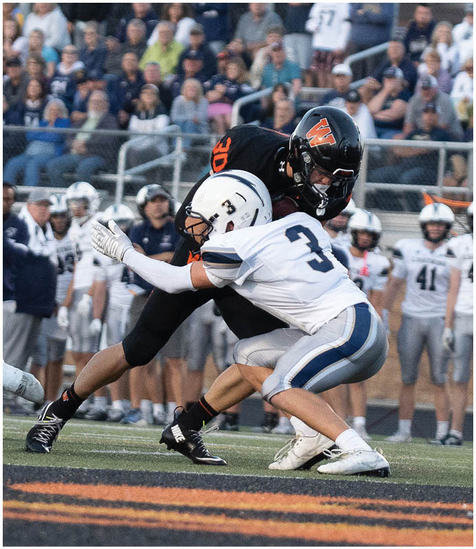 Two football players in white and black uniforms are engaged in a tackle on a field with a crowd of people watching from the stands. Description generated by AI